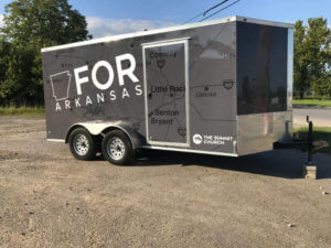 A trailer parked on gravel shows a graphic map of Arkansas with the words FOR ARKANSAS and The Summit Church on its side. Towns like Conway, Little Rock, Benton, and Bryant are visible on the map.