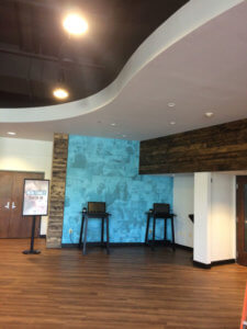 A modern lobby area with wood flooring, two check-in kiosks against a blue accent wall with faded images of people, a sign that reads “New Family Check-In,” and a mix of white and wood-paneled walls.