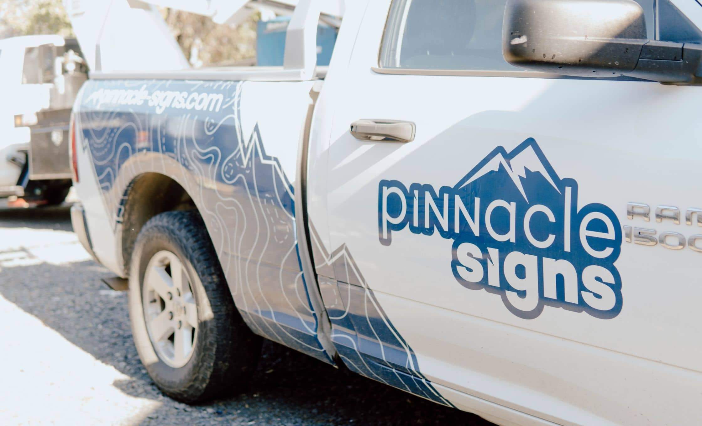 pinnacle signs truck White pickup truck with a Pinnacle Signs logo and mountain graphic on the side door. The design is blue and white, and theres a website URL on the truck bed. The vehicle is parked on a sunny day.