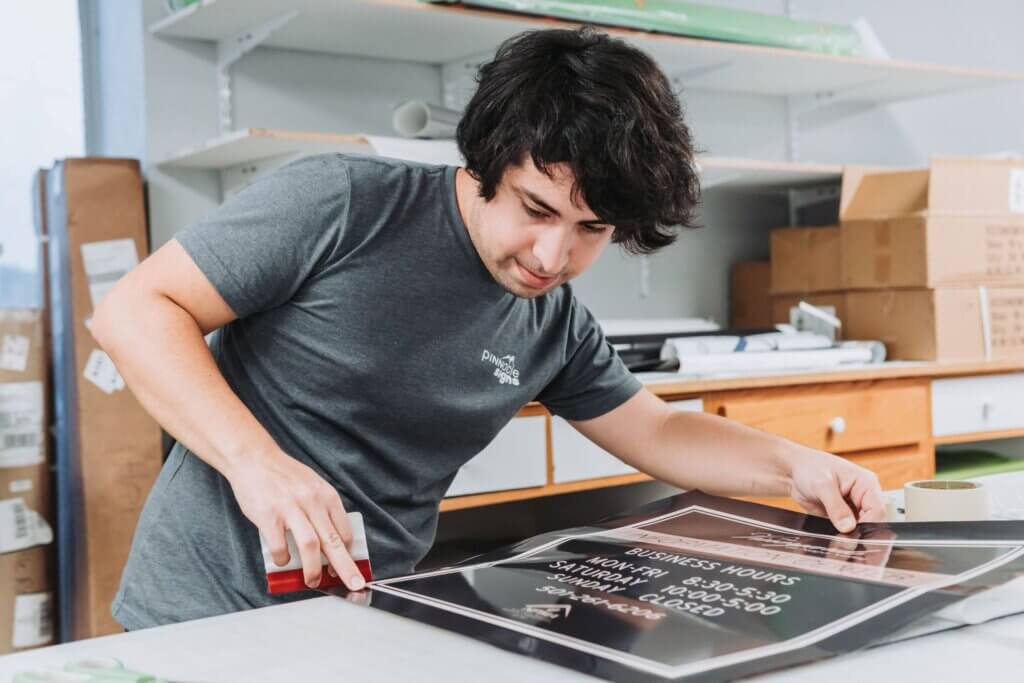 A person uses red tape to seal a framed sign featuring business hours, showcasing custom signage design skills while working at a table surrounded by shelves, boxes, and office supplies.