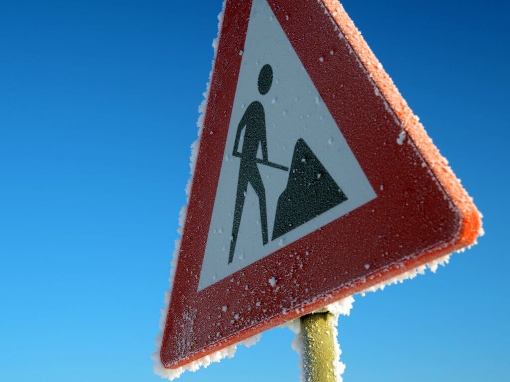 A triangular construction warning sign with frost along the edges stands against a clear blue sky, serving as effective construction signage to indicate roadwork ahead.
