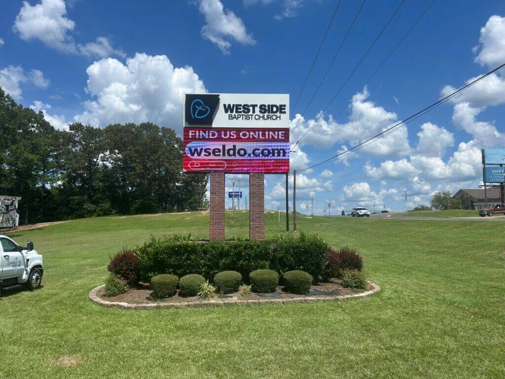 A digital sign for West Side Baptist Church displays Find us online and wseldo.com, set on a grassy area with bushes, trees, a truck, and a partly cloudy sky in the background.