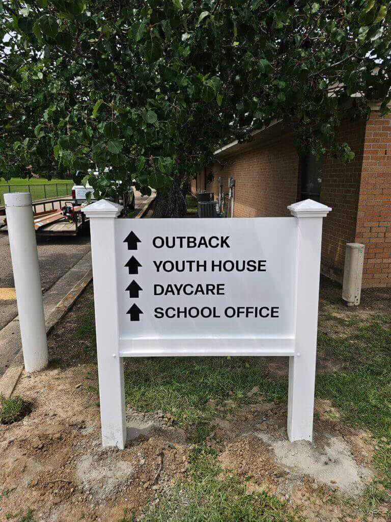 White sign with black arrows and text pointing up to Outback, Youth House, Daycare, and School Office, standing outside near a tree and a brick building.