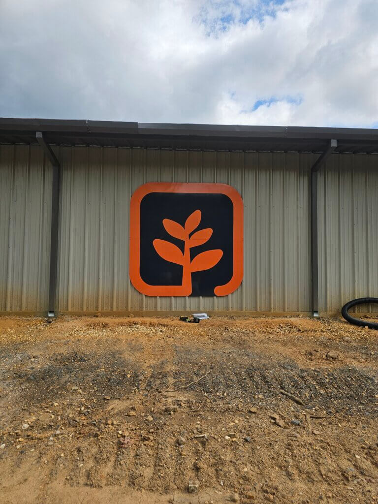 A metal building with a large orange and black sign showing a stylized plant or leaf inside a rounded square frame, mounted on the wall. Dirt and some construction materials are on the ground in front.