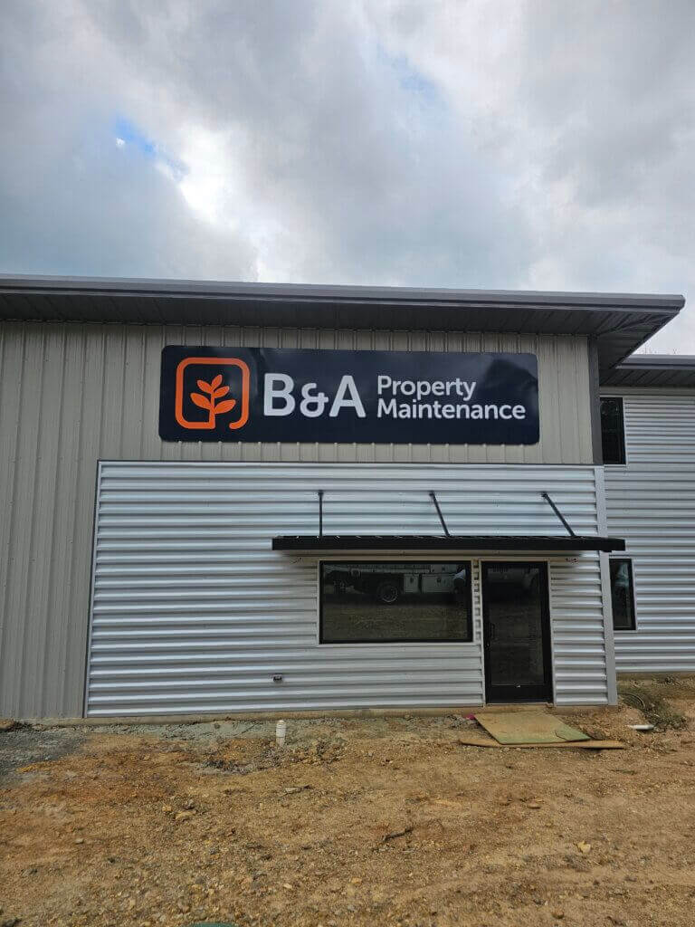 A gray metal building with a sign reading B&A Property Maintenance above the entrance. The ground in front is bare dirt and cloudy sky is visible overhead.