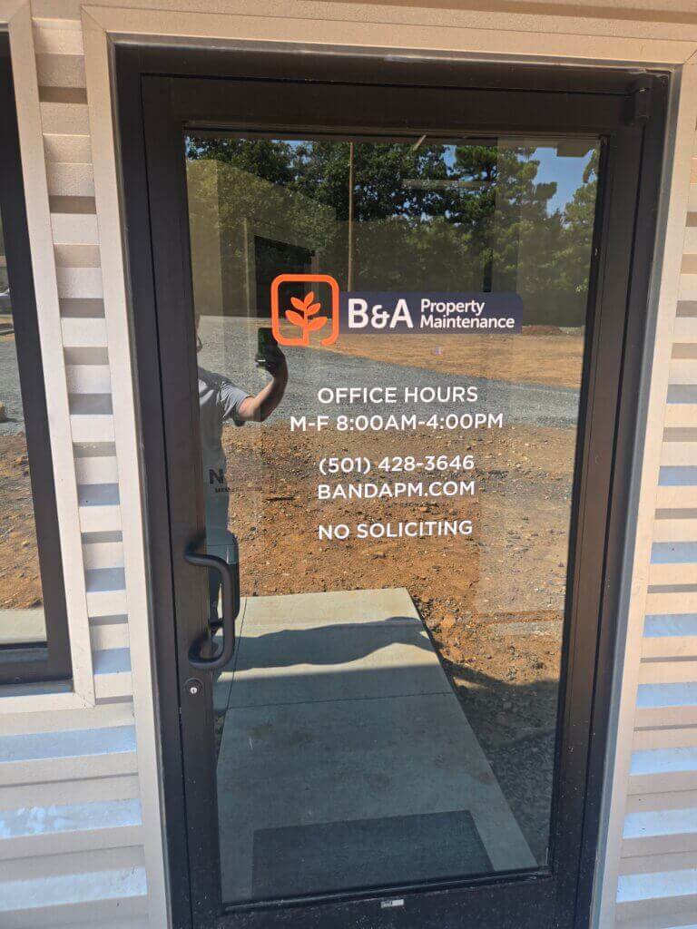 Glass door with B&A Property Maintenance logo, office hours (Mon-Fri 8AM-4PM), phone number, website, and No Soliciting text. A persons reflection and outdoor scenery are visible.