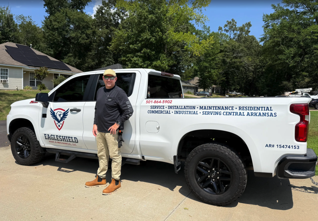 Man wearing sunglasses and a black hat standing next to a white EagleShield pickup truck parked on a suburban street. The truck displays business service information on its side. Trees and houses appear in the background.