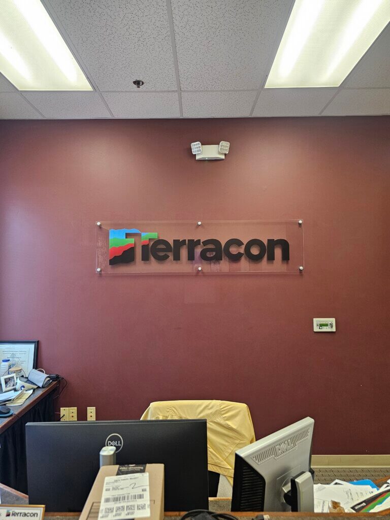 Office reception area with maroon wall, Terracon logo sign, two desks with computers, various office items, and fluorescent ceiling lights above.